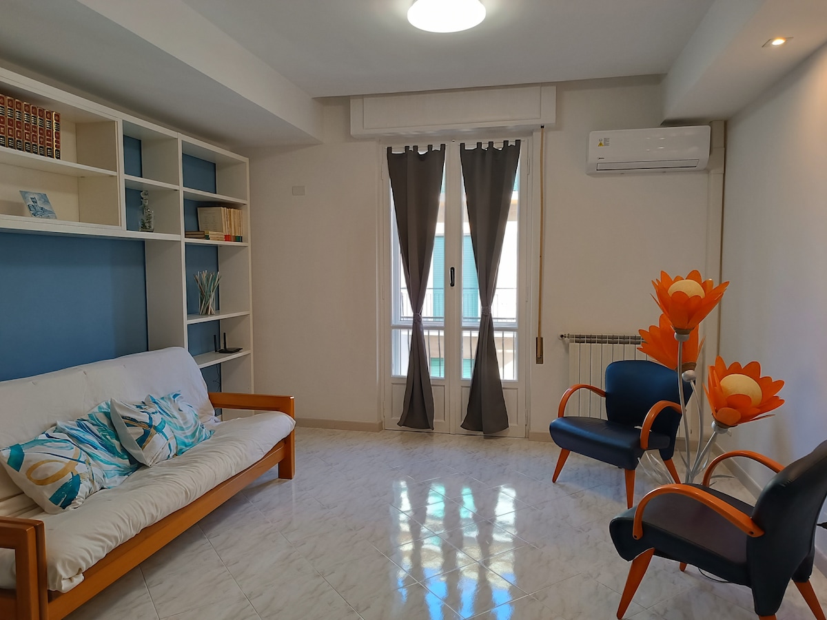 A bright living area is presented with a futon sofa bed and two armchairs, accompanied by decorative flower accents. Natural light filters through large windows covered by floor-length curtains, enhancing the spacious feel of the room. Air conditioning is conveniently positioned on one wall.