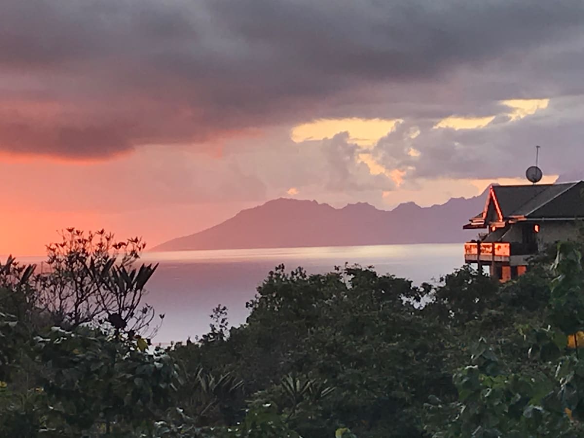 A serene view captures the vivid colors of sunset reflecting on the distant mountains and calm waters. The landscape is framed by lush vegetation, showcasing the natural beauty of the surrounding area.