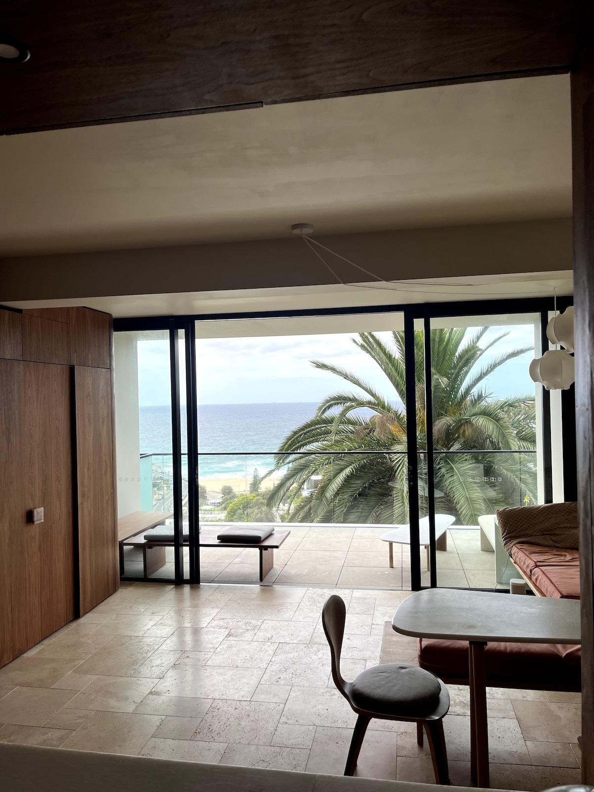 A spacious living area is showcased with floor-to-ceiling glass doors opening to a balcony with ocean views. Bright palm trees frame the scene, while a wooden seating area and a small table are present. Natural light fills the room, highlighting the minimalistic design.