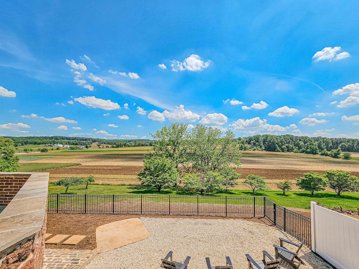 A panoramic view captures rolling farmland under a bright blue sky. The landscape features green trees and fields separated by a neat fence, with a gravel patio and seating area visible in the foreground, enhancing the outdoor space.