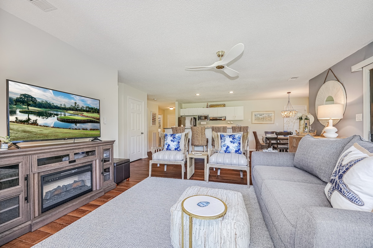 A spacious living area showcases a large flat-screen TV and comfortable seating arrangements. A light grey sofa complements two accented chairs with blue and white patterns. A neutral rug anchors the space, with a view of the kitchen and dining area in the background.