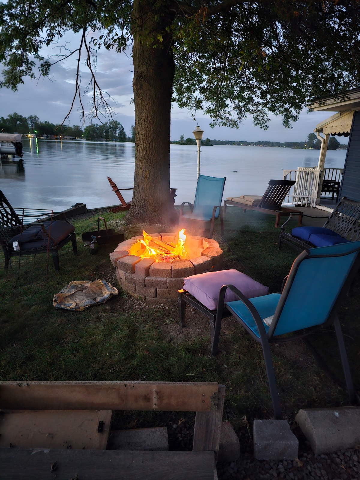 A circular fire pit made of stones is surrounded by several chairs, featuring a warm fire at its center. The serene waterfront is visible in the background, with gentle waves reflecting the evening light. Soft seating options provide comfort for guests enjoying the outdoor space.