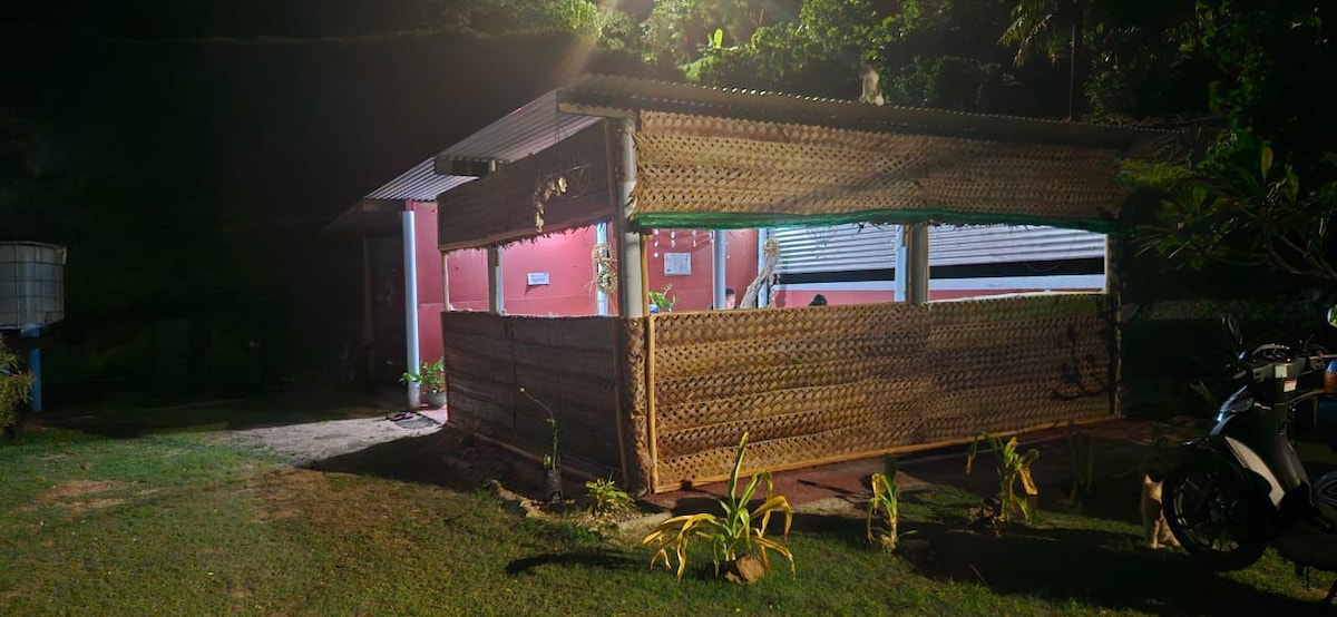 A well-constructed structure is showcased, featuring woven wall panels and an open design. The exterior is illuminated by soft lighting, revealing a surrounding grassy area with small plants. The entry is framed by a covered patio, enhancing the connection to nature.