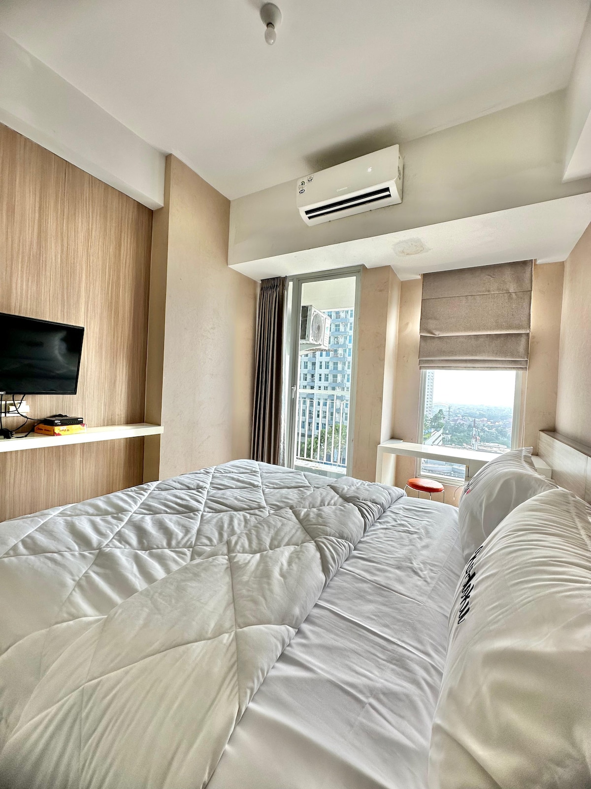 A cozy sleeping area features a spacious bed covered with a white quilt. A flat-screen TV is mounted on the wall, and natural light illuminates the space through large windows, offering a view of the city. Air conditioning is installed for comfort.