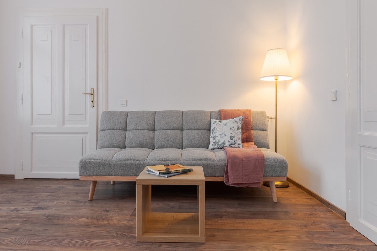 A modern gray sofa is positioned next to a wooden coffee table on a warm-toned floor. A soft throw and decorative pillow add subtle color. A floor lamp provides gentle lighting, enhancing the inviting feel of the living space.