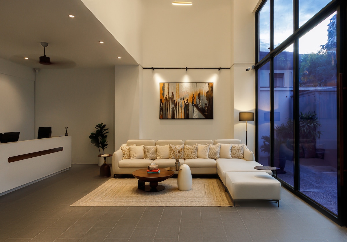The lobby area features a spacious seating arrangement with a light-colored sofa and round coffee table. A large artwork adorns the wall, while large windows provide natural light and views of the outside. A reception desk is visible in the background, enhancing the welcoming environment.