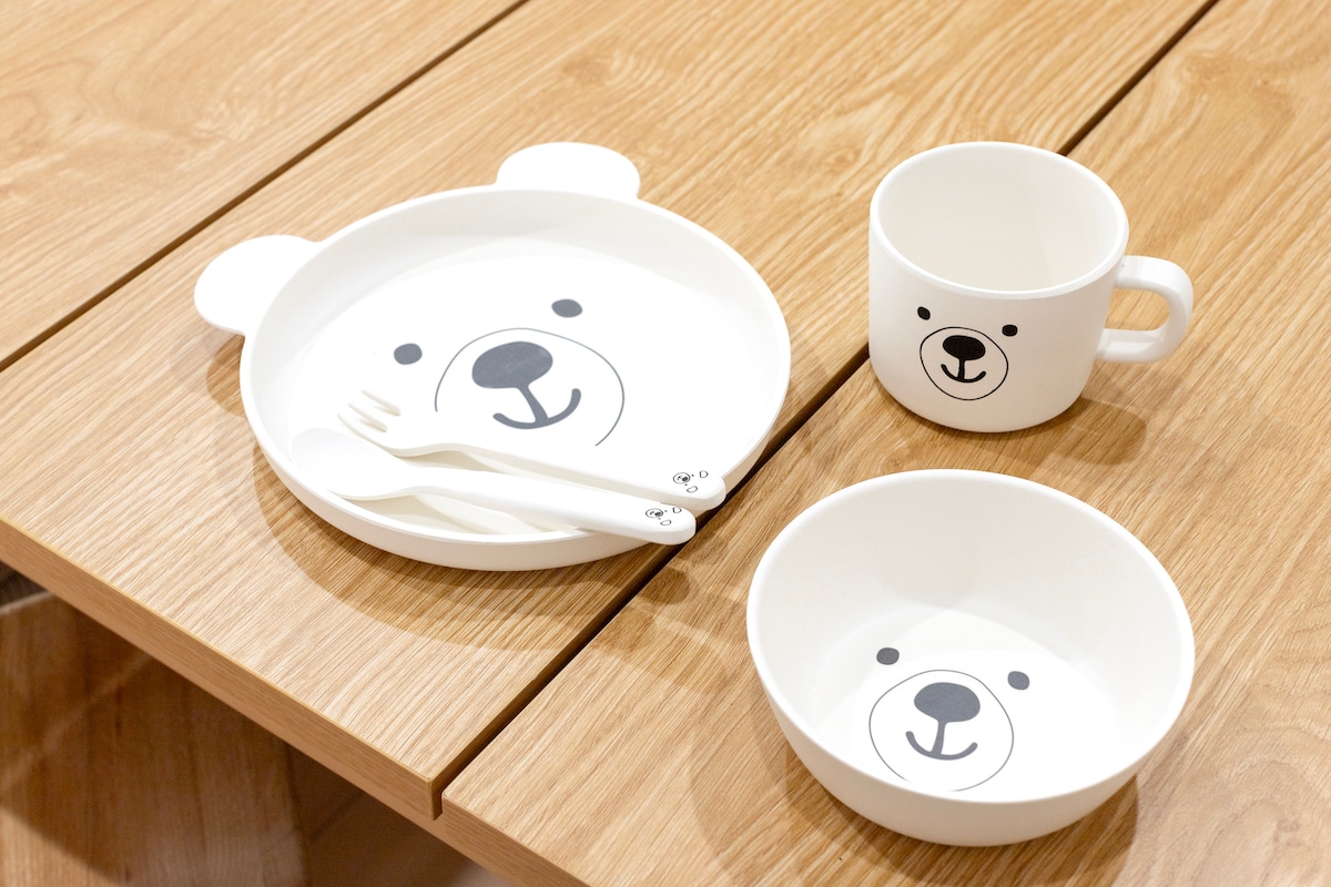A playful set of children’s tableware is displayed on a wooden table, featuring a bowl, plate, and cup, each adorned with a bear face design. A fork and spoon accompany the plate, providing a cheerful dining experience.