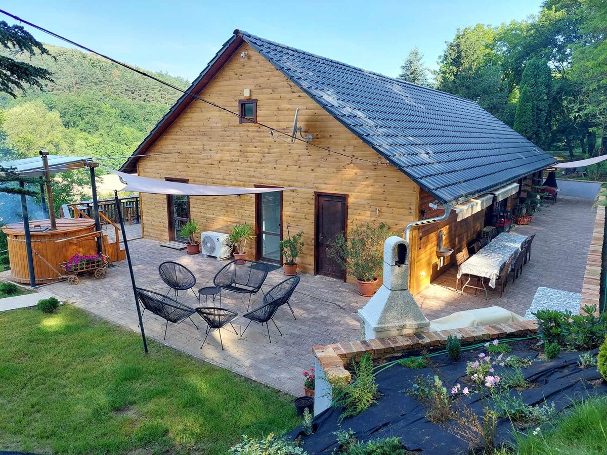 A charming wooden guesthouse is surrounded by lush greenery, featuring a spacious outdoor patio area. Comfortable seating arrangements and a long dining table are positioned for gatherings. A wooden hot tub and sauna are visible nearby, enhancing the serene atmosphere of the property.