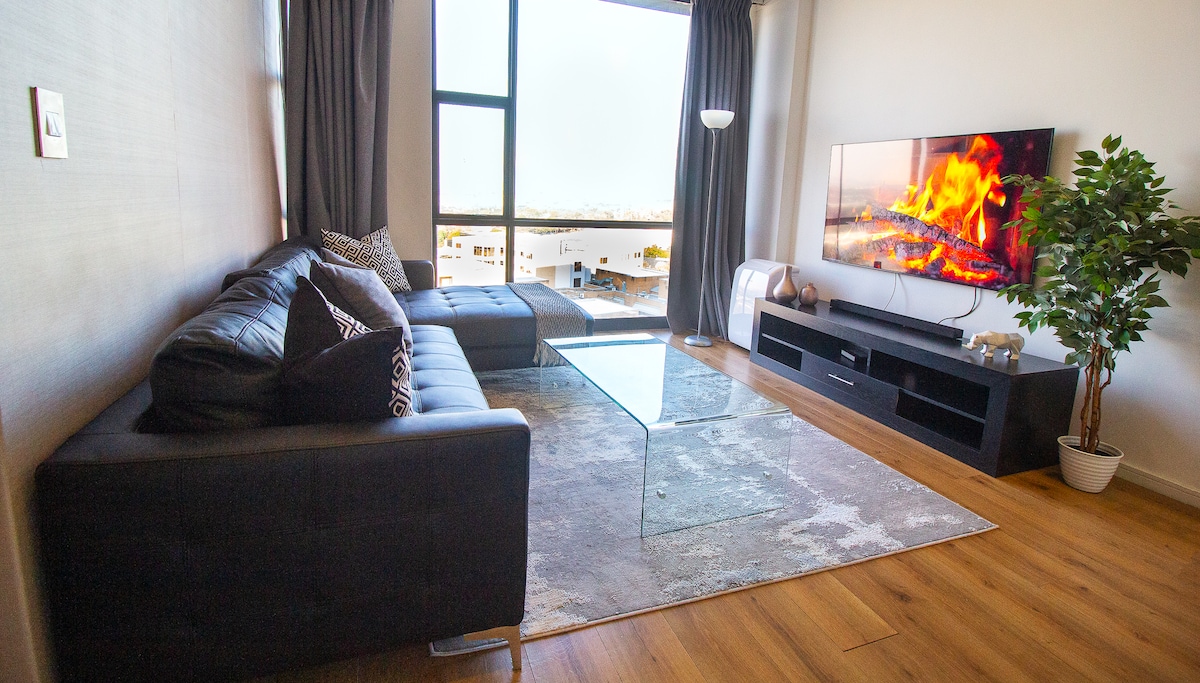 A stylish living area features a dark leather sectional couch and a modern glass coffee table. A large television displays a warm fire scene, while natural light floods in through the floor-to-ceiling window. A small plant adds a touch of greenery.