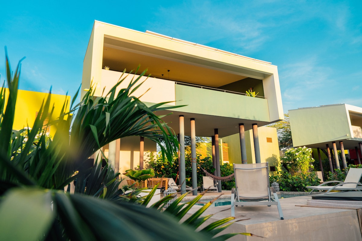 The villa features a contemporary architectural design with two levels and a clean, modern façade. Lush greenery surrounds the property, and outdoor lounge chairs are positioned on the terrace, inviting relaxation under the bright, sunny sky.