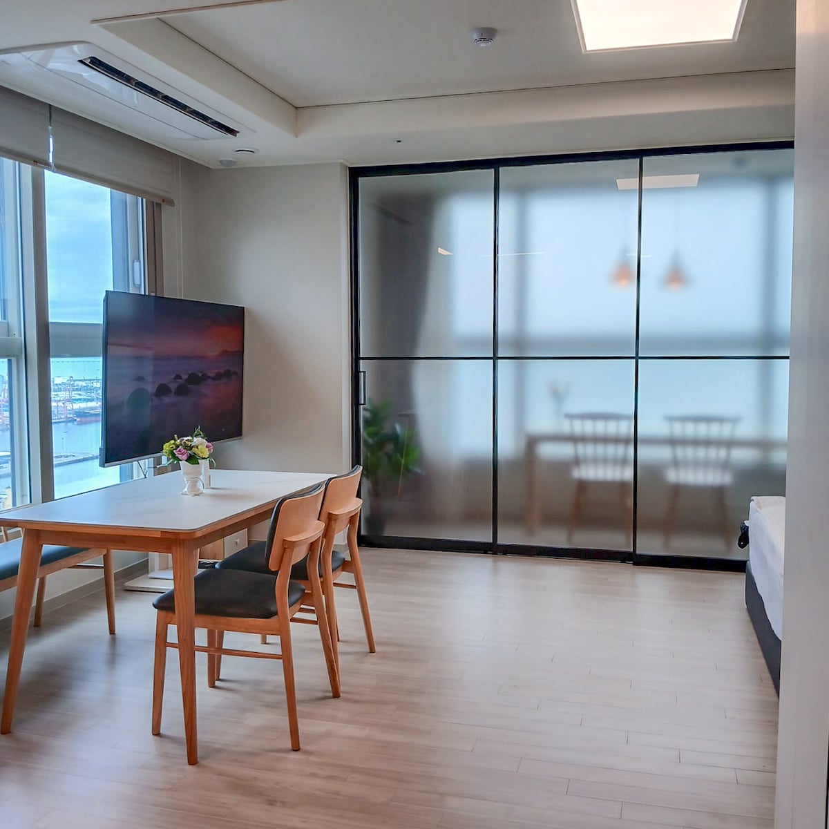 A bright living area features a dining table set for four, complemented by simple wooden chairs. A large television stands against the wall, and soft natural light enters through large windows. A glass partition separates the space, creating a gentle division with an unobstructed view.