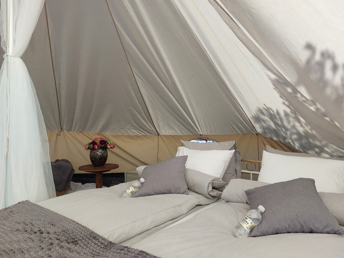 The interior of the spacious tent features a comfortable sleeping area with layered bedding and decorative pillows. A small table holds a vase with flowers, and two bottled waters are placed on the bed. Soft, muted colors create a calming ambiance.