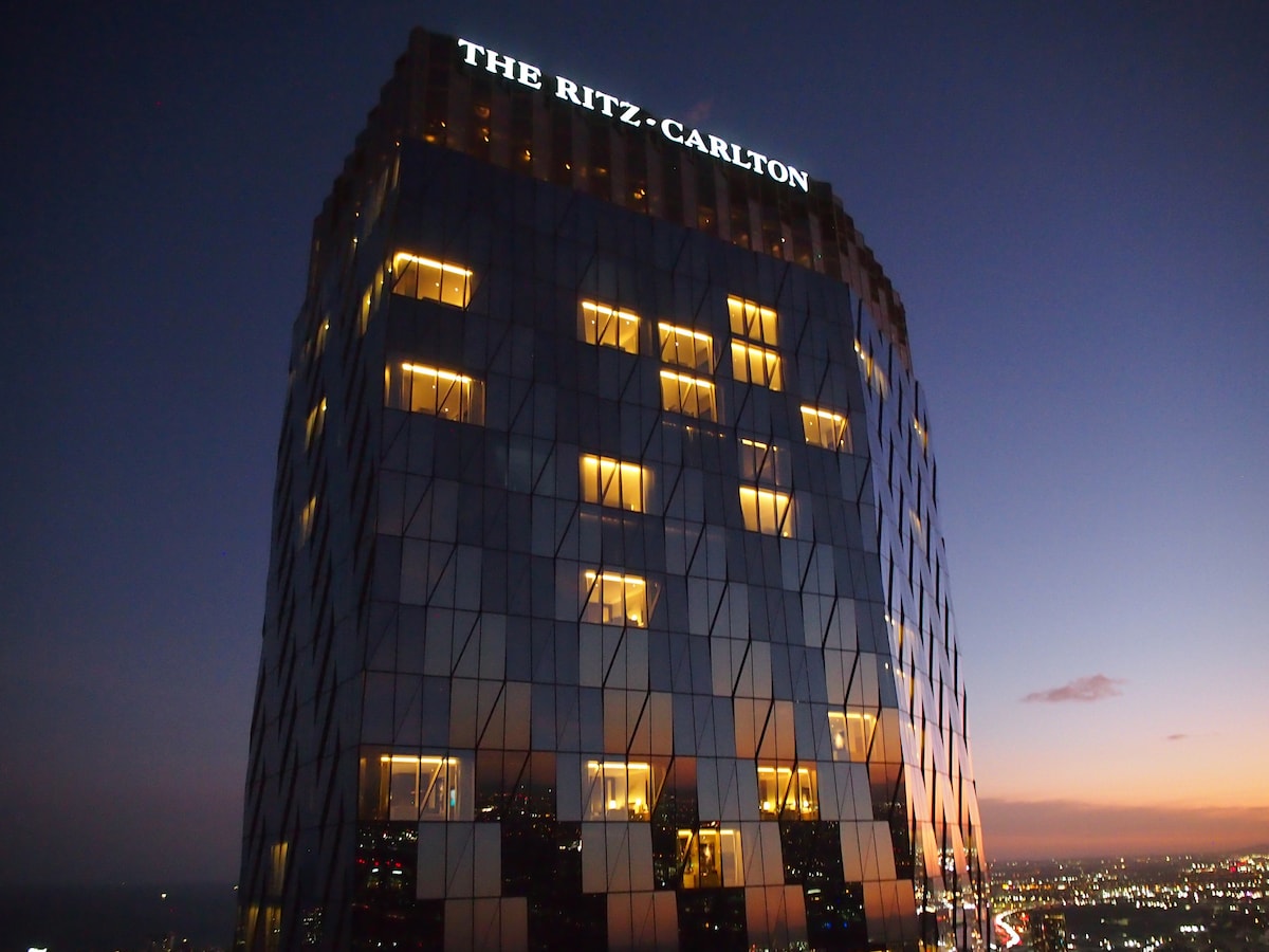 The illuminated façade of The Ritz-Carlton at dusk is presented, showcasing its modern architectural design. The building features reflective glass showcasing warm lights from the interior. A gradient sky transitions from twilight to night, creating a serene backdrop.
