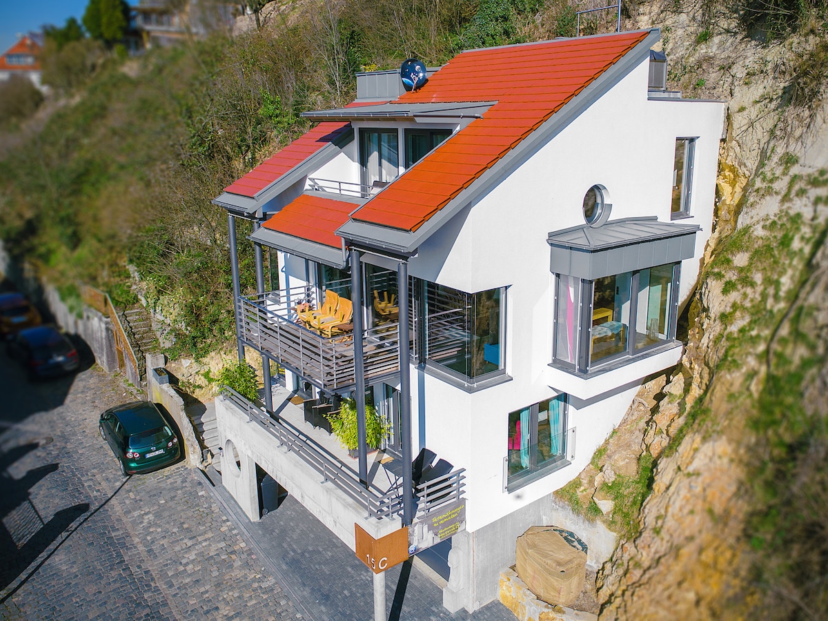 The modern, multi-level building features a striking red sloped roof and large glass windows. A spacious balcony is adorned with wooden seating, overlooking the surrounding greenery. The structure is positioned on a hillside, providing a unique architectural presence in the landscape.