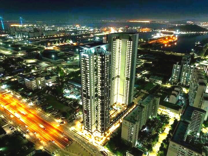 The image captures a panoramic view of a modern high-rise building at night, illuminated by city lights. The surrounding urban landscape features a mixture of residential and commercial structures, with the glow of traffic visible on the busy road below.