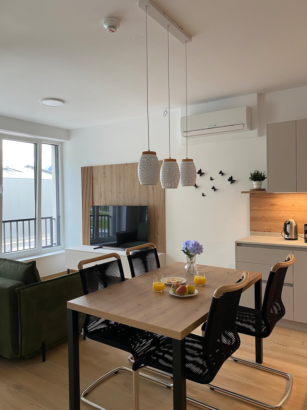 A dining area visible in the image features a wooden table surrounded by four black chairs. Above the table, three pendant lights hang, while a small bowl of fruit and two glasses of juice are placed on the table. Natural light enters through the large window, illuminating the modern kitchen in the background.