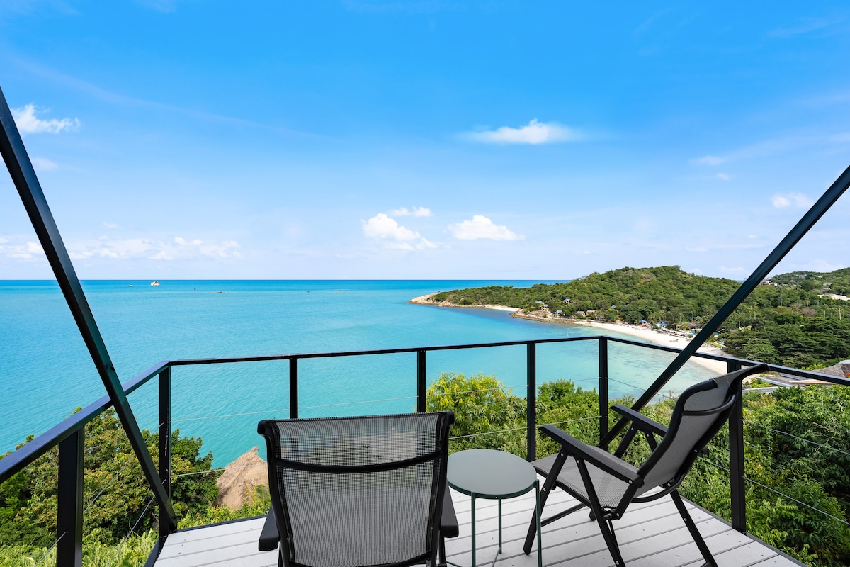 A patio area features two black lounge chairs alongside a small round table, overlooking a breathtaking view of the turquoise sea and nearby coastline. The glass railing allows unobstructed sight of the water and natural surroundings under a clear blue sky.