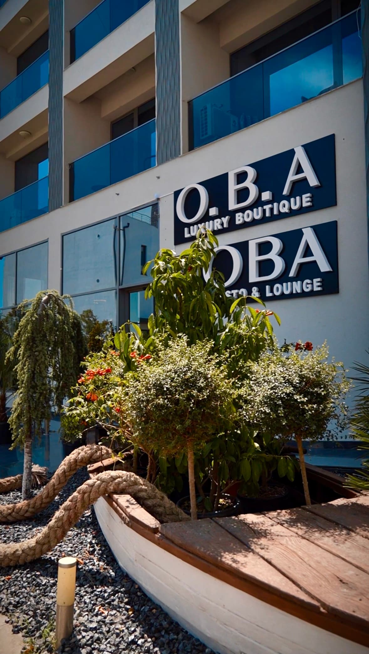 The exterior of a luxury boutique is shown, featuring a prominent sign displaying 'O.B.A. Luxury Boutique.' Surrounding greenery includes potted plants and large trees. A wooden boat with rope accents is positioned in the foreground, along with decorative gravel pathways.