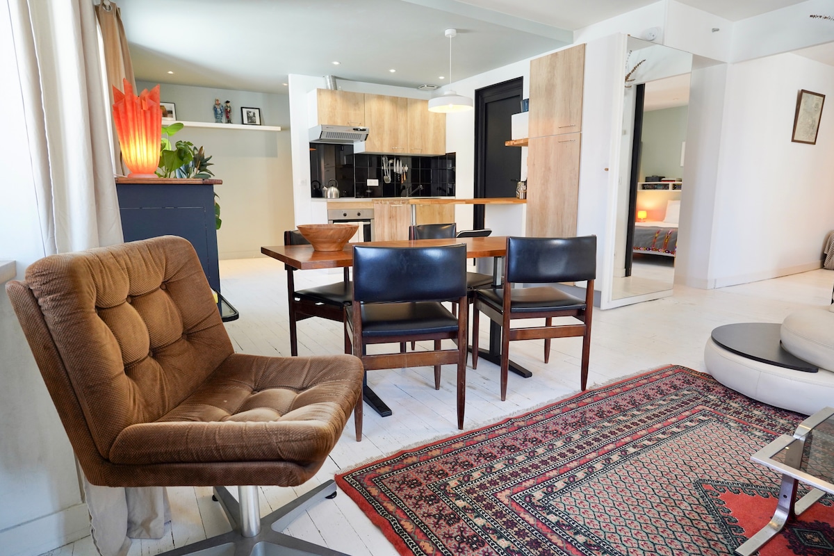 The living area features a contemporary kitchen with wooden cabinetry and sleek appliances, open to a dining space, which includes a table for four. A comfortable chair is positioned near the rug, while light enters through large windows, illuminating the overall space.