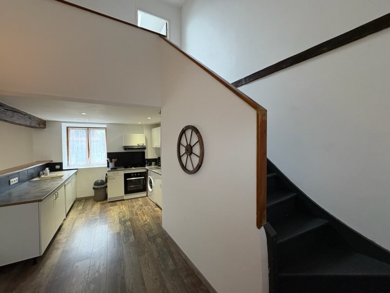 A modern kitchen is seen with light cabinetry and a dark countertop, accompanied by a stove, oven, and washing machine. A staircase with wooden railing leads to an upper level. Natural light enters through a nearby window, illuminating the space's clean lines.