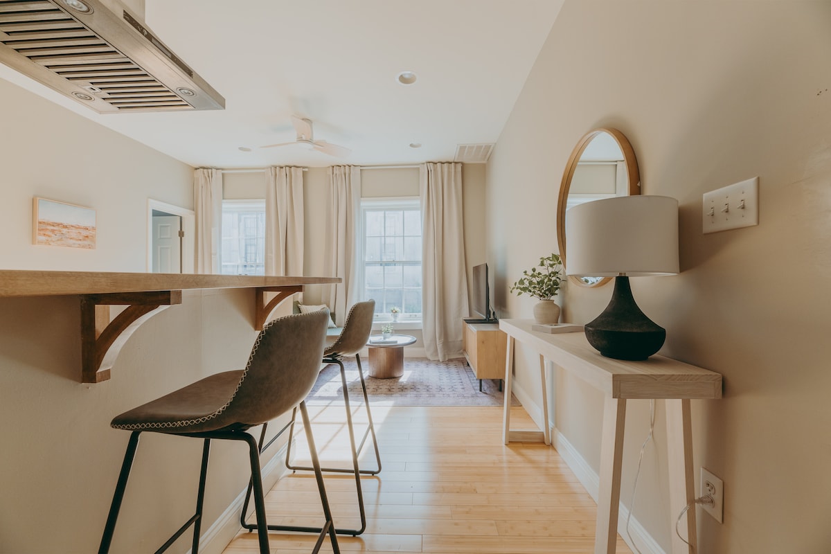 A modern kitchenette area with a wooden bar counter and two stylish stools is visible. Natural light floods the space through large windows, highlighting soft beige walls and warm wood flooring. In the background, a cozy living area is seen with a coffee table, television, and decorative elements.