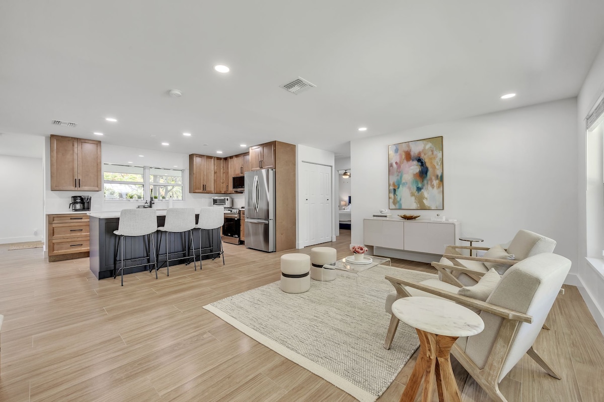 An open-concept living area features a modern kitchen with wood cabinetry and stainless steel appliances. A dining area with bar stools offers a casual eating space. Comfortably arranged seating, including light armchairs and a neutral area rug, creates an inviting space with natural light from large windows.