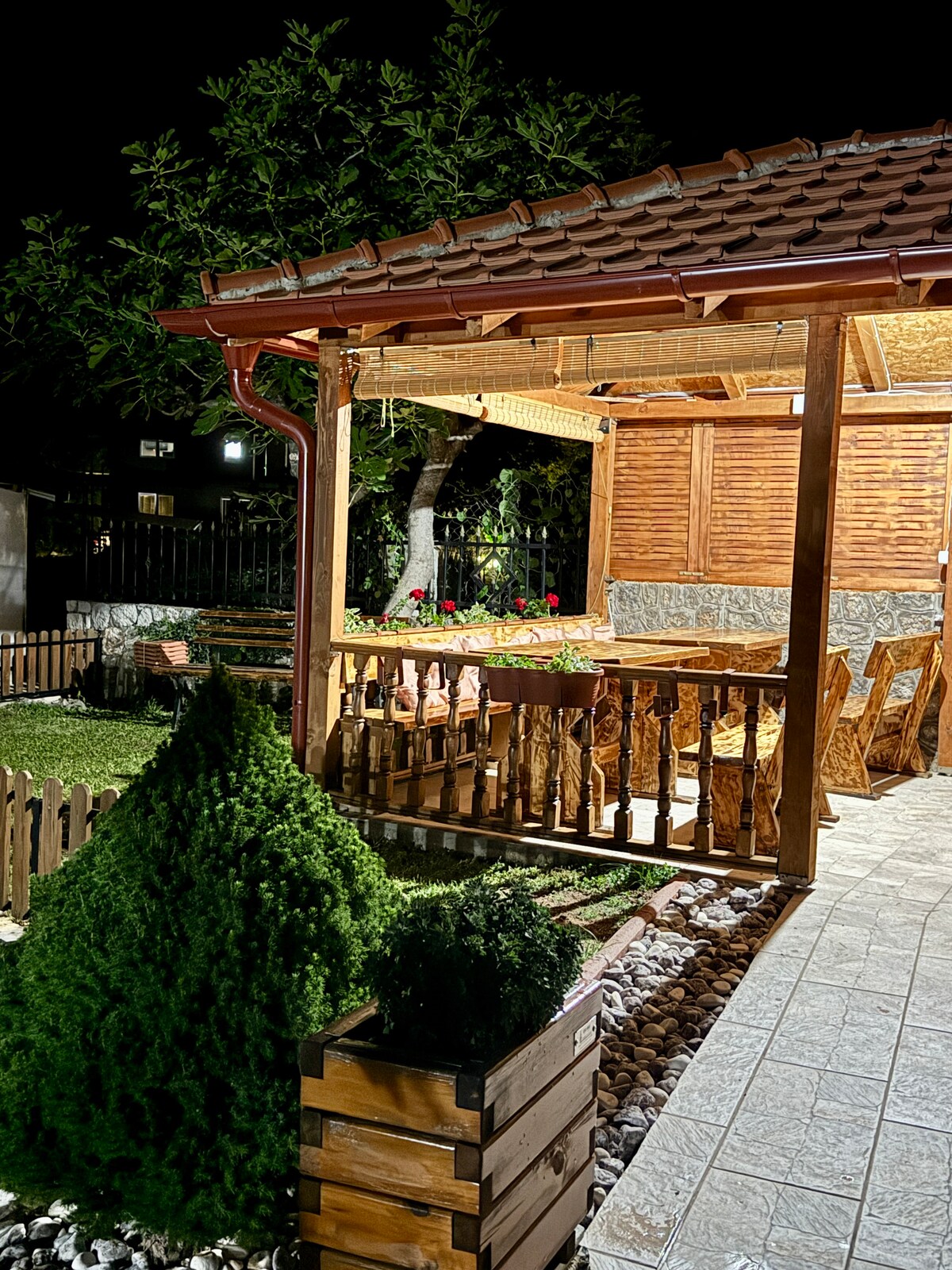 An outdoor dining area is illuminated at night, featuring wooden furniture under a covered structure. A garden with manicured shrubs and decorative stones surrounds the space, enhancing the inviting atmosphere. Soft lighting highlights the textures of the wood and the greenery.