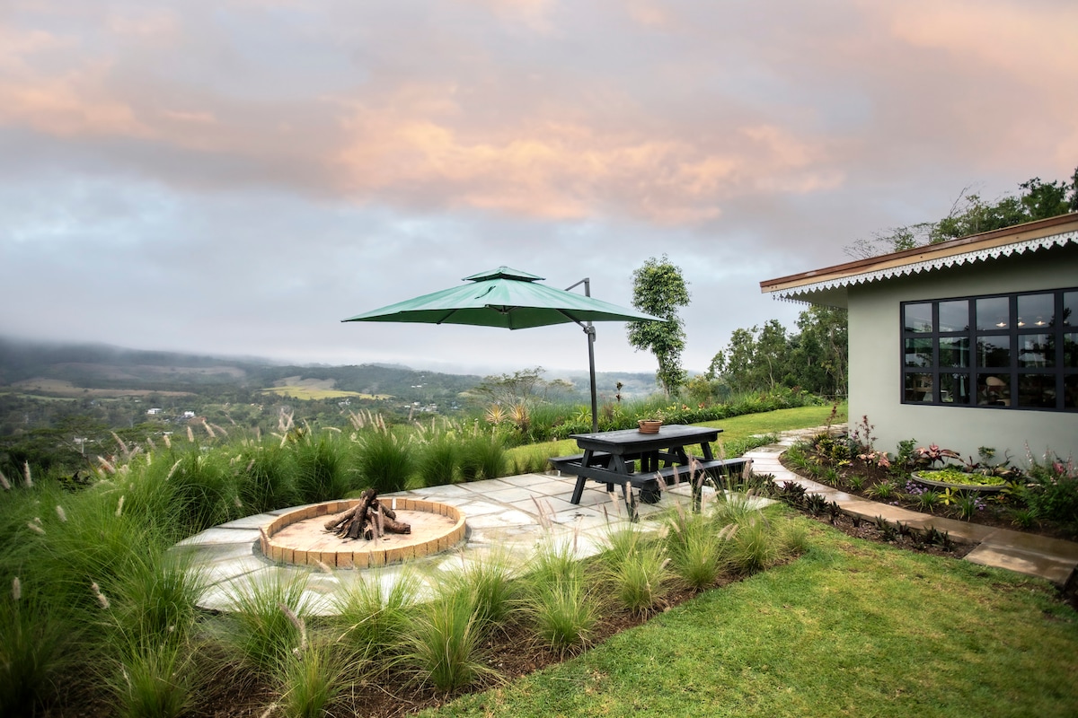 A patio area features a circular fire pit surrounded by low stone seating, complemented by a wooden picnic table beneath a large green umbrella. Lush grass and ornamental plants frame the scene, with a panoramic view of rolling hills and a cloudy sky in the background.