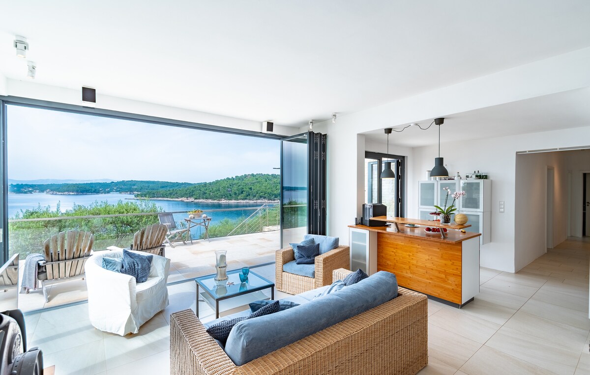 A spacious living area features large glass doors that open to a terrace, providing a view of the surrounding bay. Comfortable seating options, including a wicker sofa and chairs, are arranged around a glass coffee table. Natural light fills the room, enhancing the open feel.