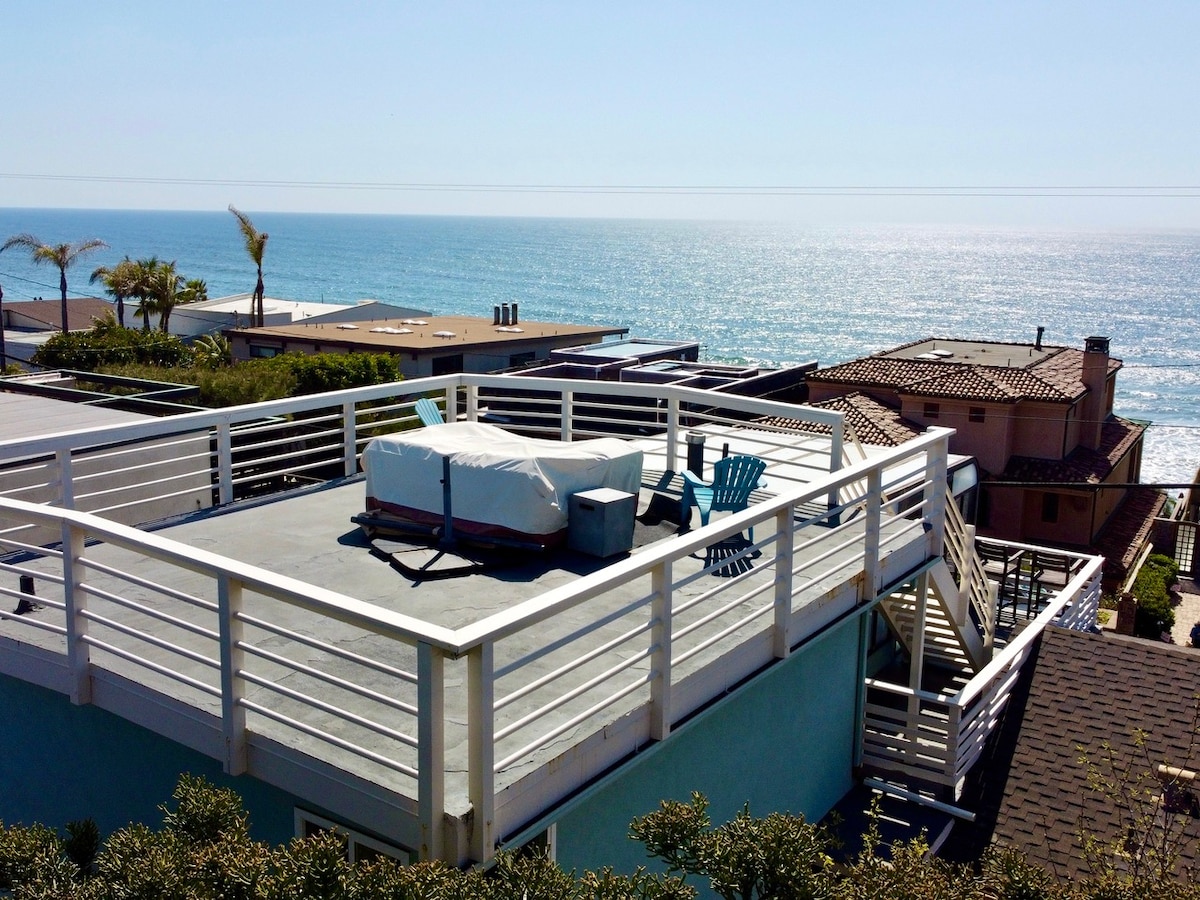 The rooftop deck offers a panoramic view of the ocean, capturing the shimmering water and coastal landscape. A white hot tub is positioned centrally, surrounded by colorful chairs for relaxation. The area is framed by wooden railings, enhancing the inviting outdoor space.