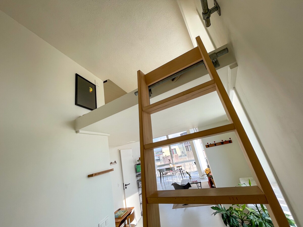 A wooden ladder leads to a sleeping platform above, highlighted by the spacious, light-filled interior. Large windows allow natural light to fill the space, creating a bright and airy atmosphere. A glimpse of the kitchen area and living space is visible below.