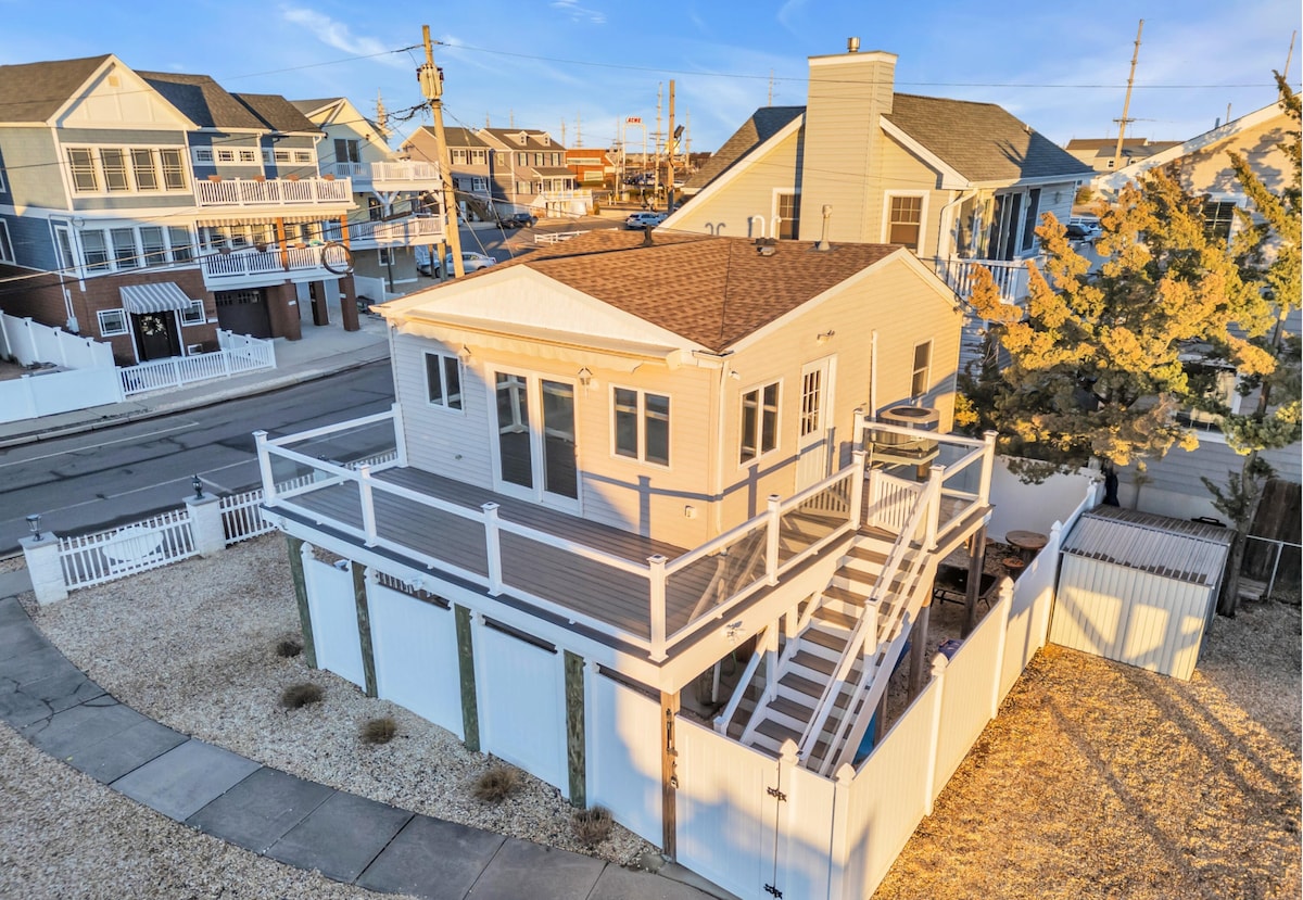 An aerial view of a charming cottage is displayed, showcasing a spacious deck surrounded by a white railing. The property is enclosed by a fence and features a landscaped yard with a gravel surface. Nearby structures reflect the coastal style typical of the area.