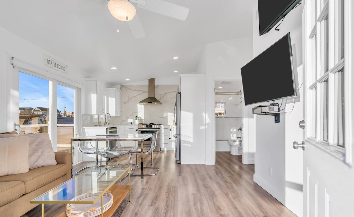 An open concept living area features a comfortable sofa paired with a sleek coffee table. Modern stainless steel appliances are visible in the kitchen. Large windows allow natural light to illuminate the space, highlighting the contemporary design with wood-like flooring and minimalistic decor.