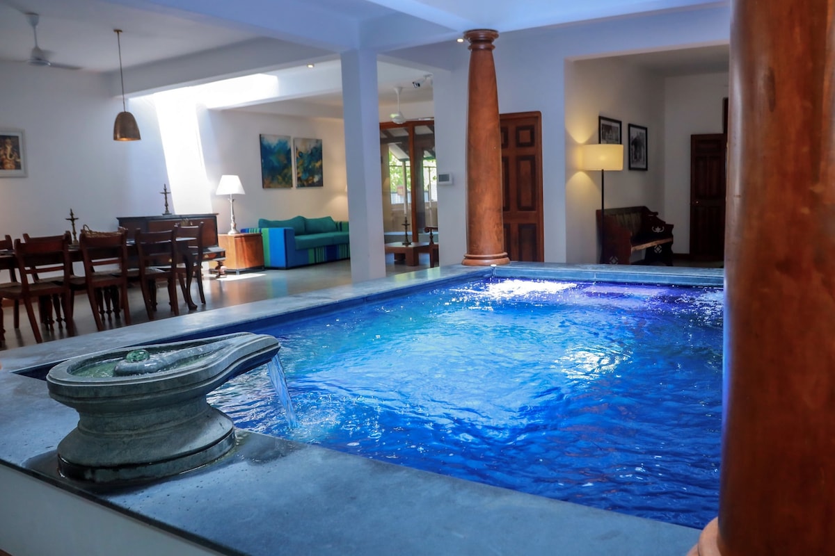 An indoor plunge pool with clear blue water is the focal point, framed by elegant columns. Natural light floods the space, highlighting the serene setting. A dining area is visible in the background, featuring a wooden table and chairs, creating a welcoming contrast.
