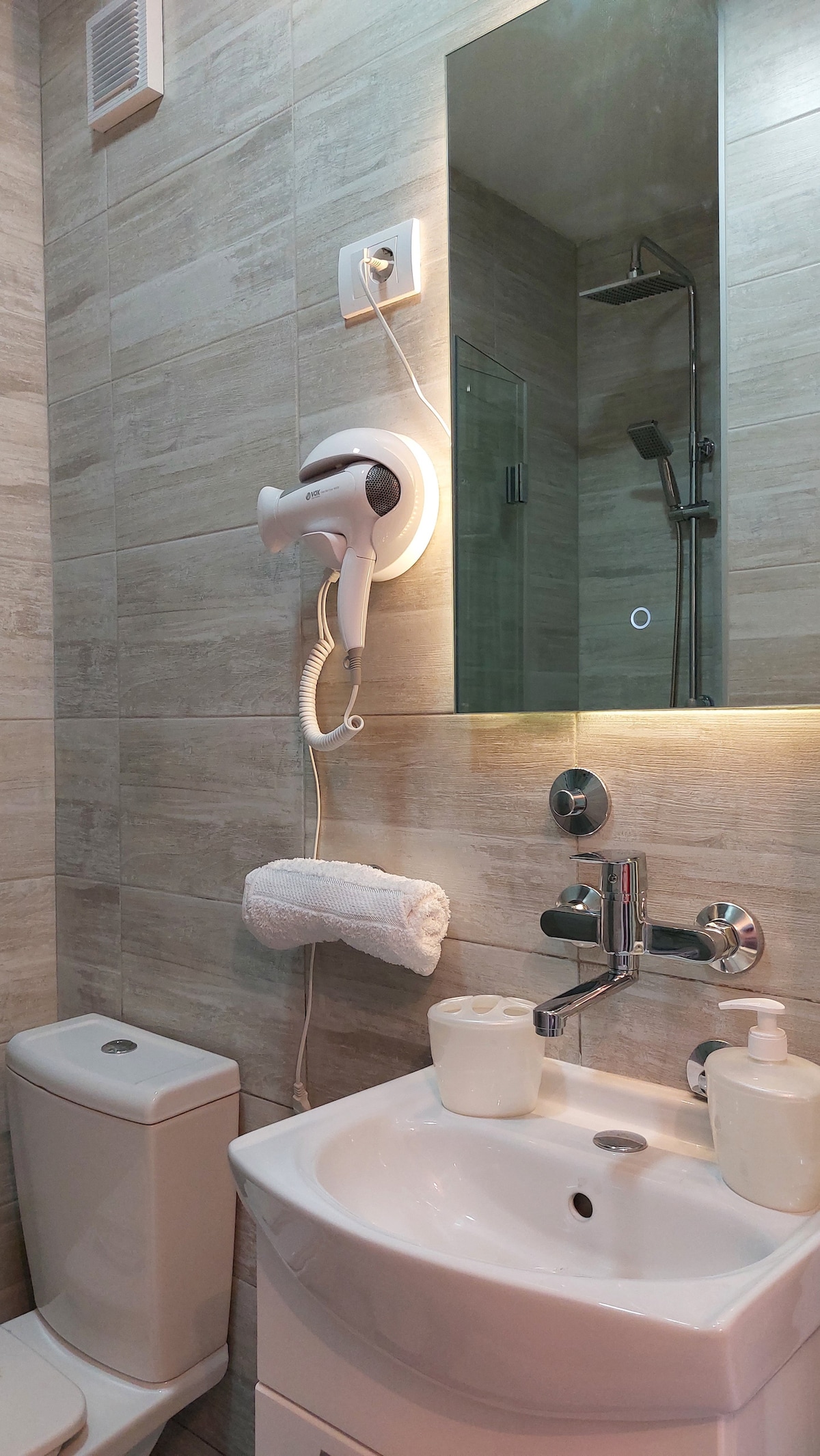 A modern bathroom is equipped with a wall-mounted hairdryer next to a mirror. A compact vanity with a sink hosts soap and toiletries. Warm lighting highlights the tiled walls, and a shower area is visible through a glass partition.
