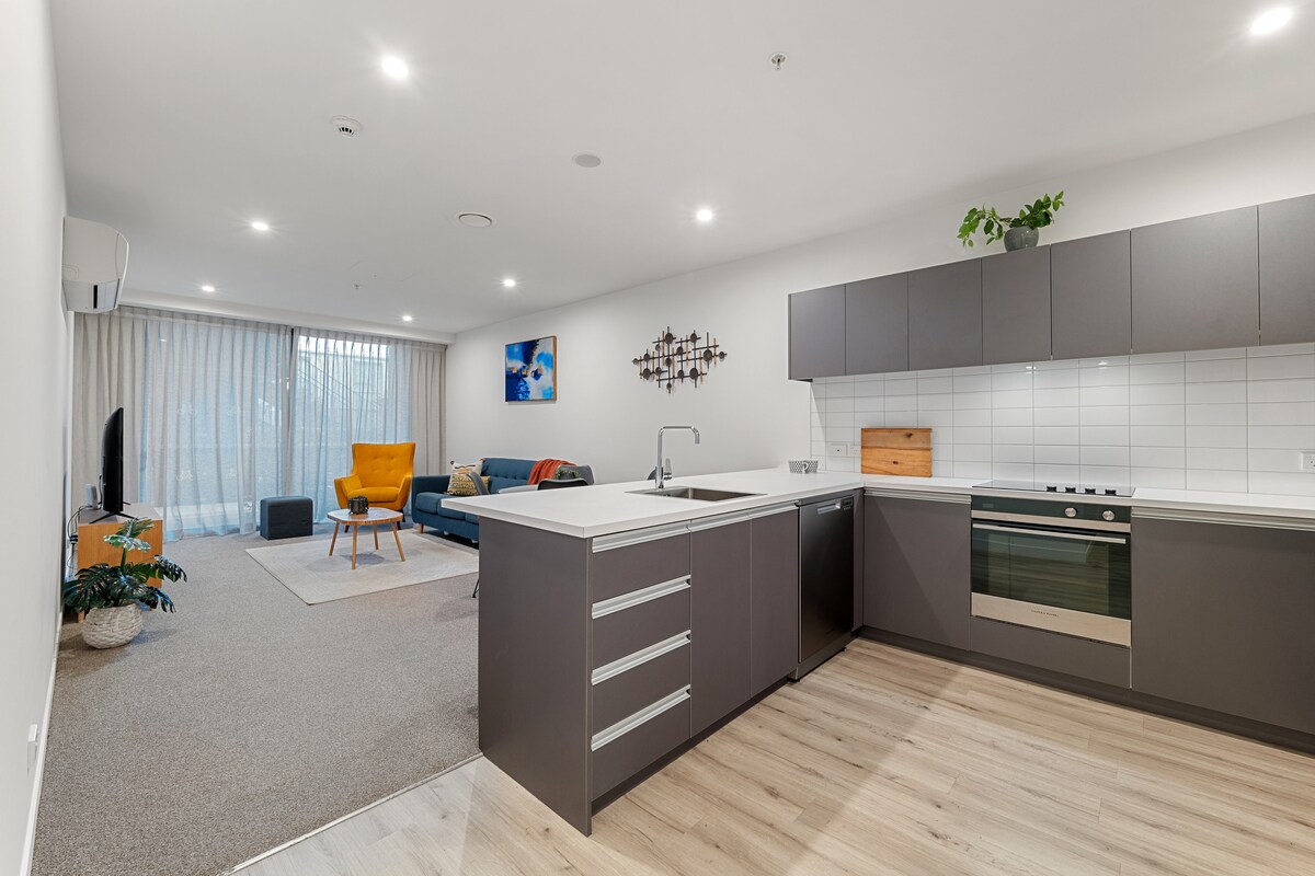 A spacious apartment interior is presented, featuring a modern kitchen with a large island and built-in oven. A cozy living area is visible, furnished with a sofa and an armchair, while natural light flows through large windows, illuminating the inviting space.