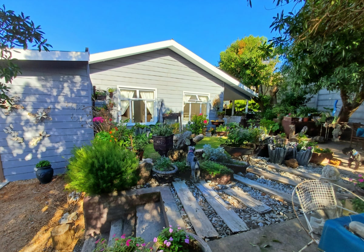 A light grey cottage is surrounded by a lush garden, filled with various plants and flowers. Pathways made of stones and gravel lead toward the entrance, while a cozy outdoor seating area is visible nearby, inviting relaxation in a serene natural setting.