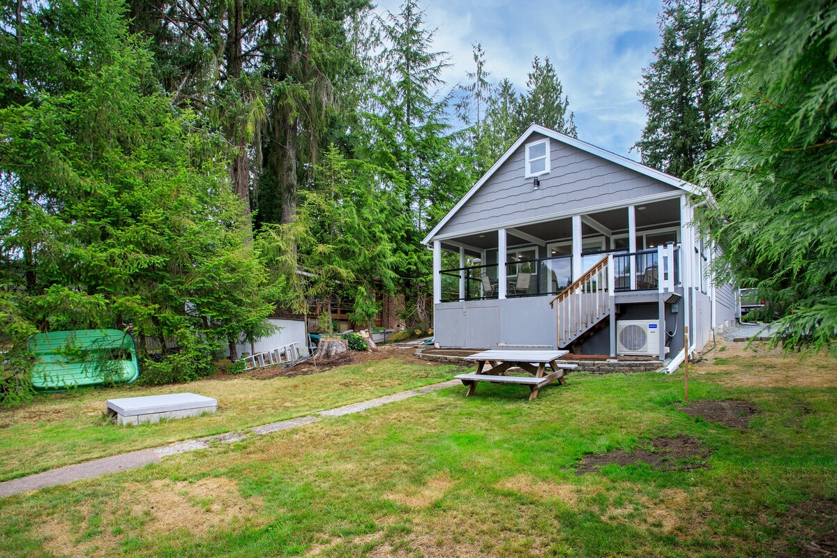 The lakeside cabin is framed by lush greenery, showcasing a spacious yard. A wooden deck with steps leads to the entrance, while picnic seating is visible on the grass. Nearby, a covered outdoor area offers a shaded space for relaxation.