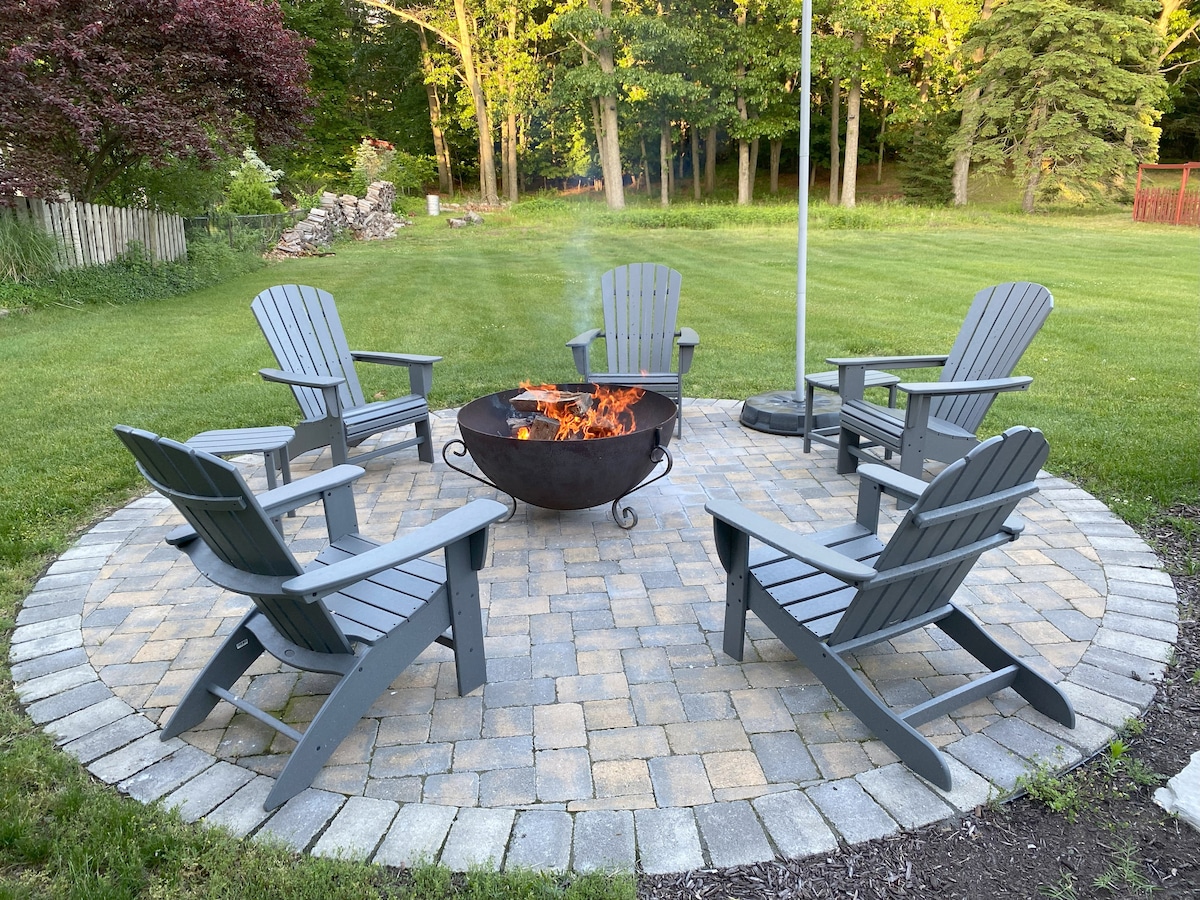 A circular fire pit area is surrounded by five spacious, gray Adirondack chairs. The stone-paved ground complements the serene green lawn and lush trees in the background, creating a peaceful outdoor gathering space for relaxation and socializing.
