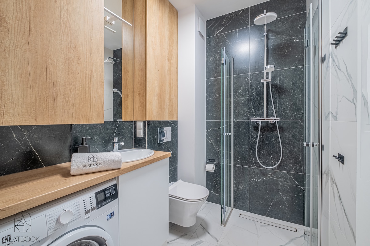 A modern bathroom features sleek dark stone walls and light wood cabinetry. A glass-enclosed shower is visible, alongside a vanity with a mirror. A washing machine is integrated into the cabinetry, offering convenience within the space.