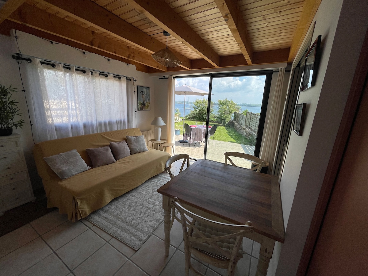 A light-filled living area features a comfortable sofa, a wooden dining table, and a view of the outdoor terrace. Large sliding glass doors open to the garden and sea, while soft drapes add texture to the bright space.