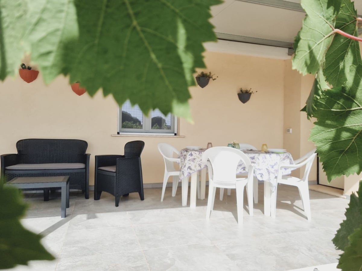 A covered patio area is visible, featuring a table and white chairs surrounded by potted plants. Comfortable seating is provided by dark wicker chairs and a low black table, offering a space for relaxed outdoor dining.