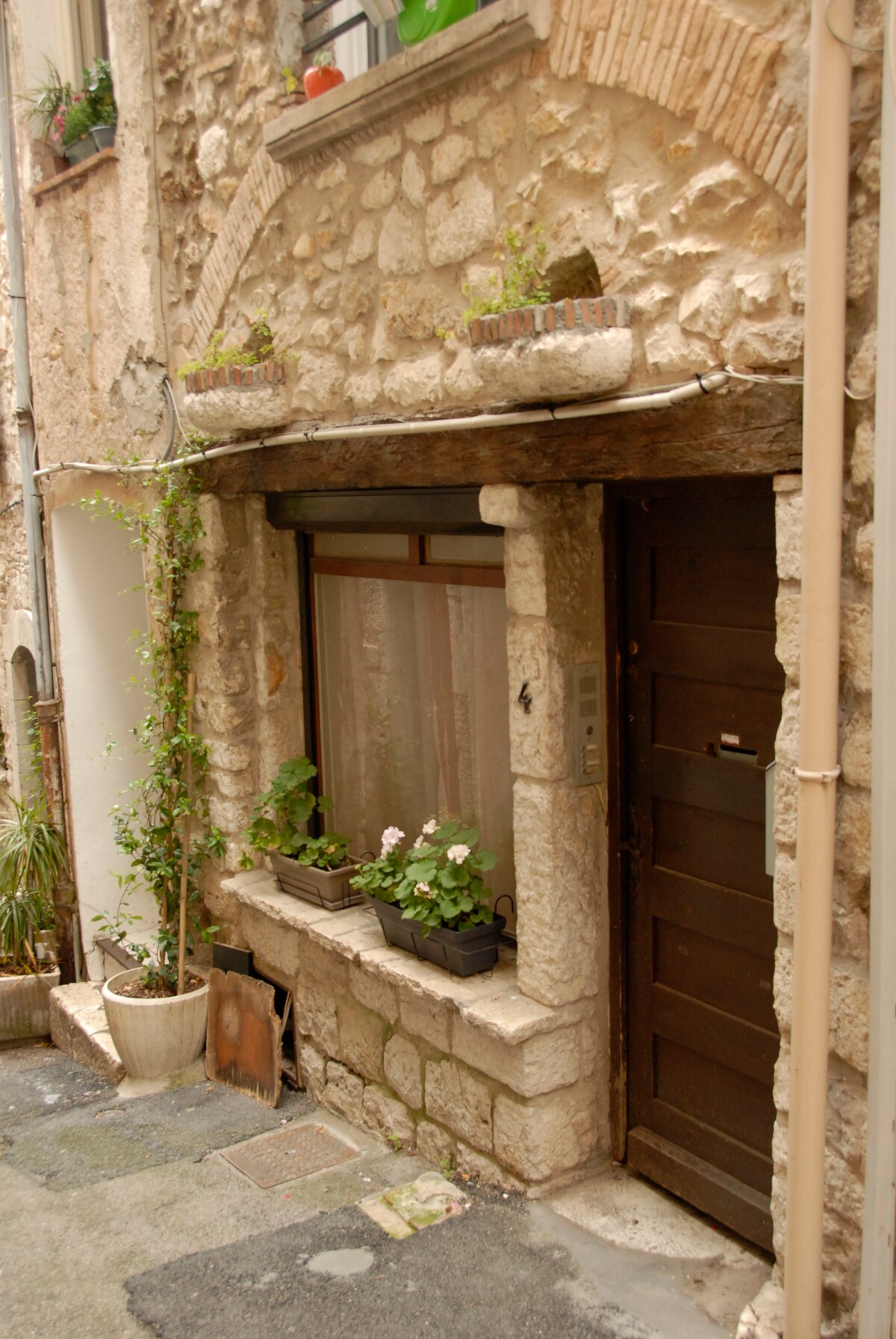A charming stone facade is featured, with an arched entryway and a wooden door. Flowering plants are placed in window boxes along the ledge, and additional greenery is seen in a nearby pot, enhancing the rustic village atmosphere.