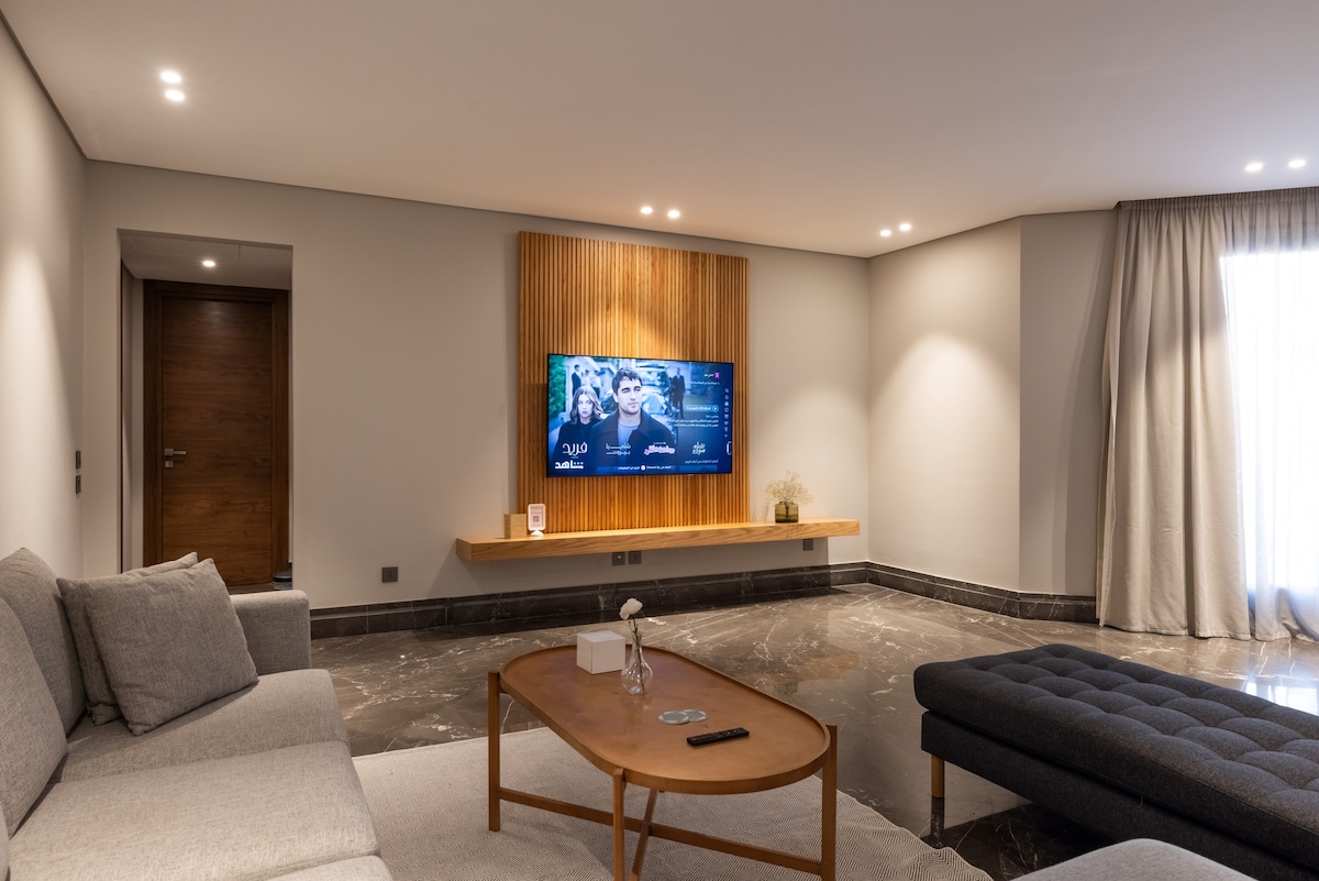 A modern living area features a comfortable gray sofa and a plush dark ottoman. A wooden entertainment unit showcases a television mounted on the wall. Soft lighting highlights the stylish decor, while large windows allow natural light to fill the space.