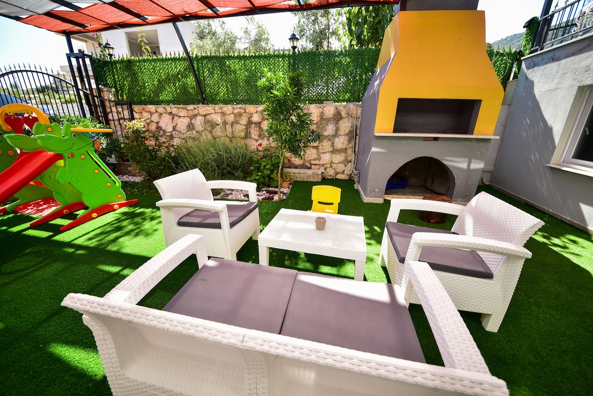 A cozy outdoor seating area is featured, with four white wicker chairs surrounding a small table on green artificial grass. A grill is visible in the corner, alongside playful garden elements, including a colorful slide. Natural light enhances the inviting atmosphere.
