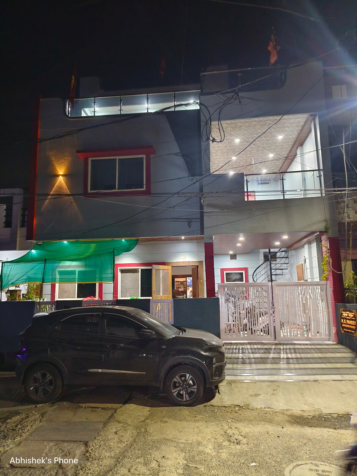 The exterior of the property is illuminated at night, showcasing a two-story structure with red accents. Large windows and a balcony are visible, surrounded by well-maintained greenery. A modern car is parked in front, and the entrance is framed by a decorative gate.