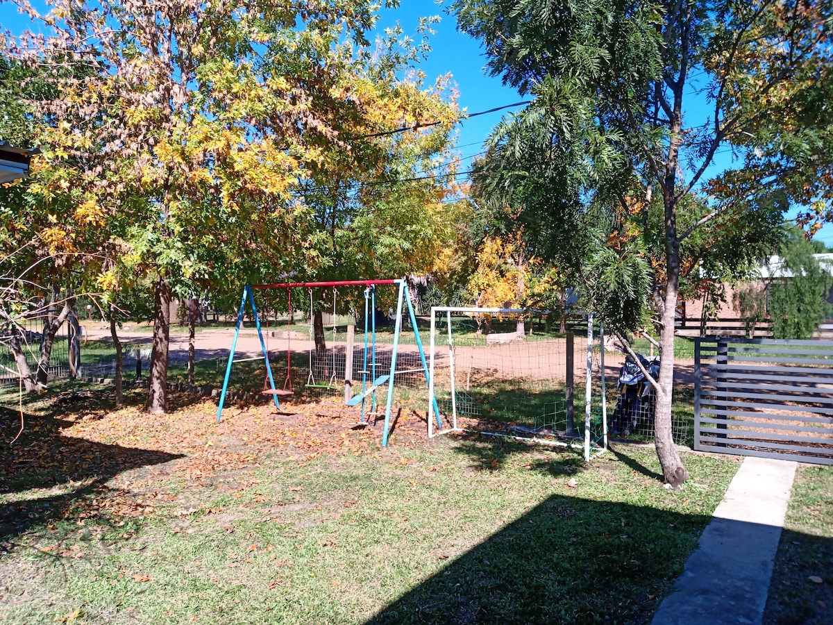 An outdoor play area features swings and a slide surrounded by trees with autumn foliage. The grassy ground provides a safe space for children to play. A fence is visible in the background, ensuring a secure environment.