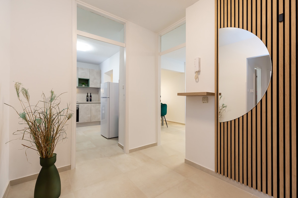 A bright entrance area is presented, featuring smooth tile flooring and neutral-colored walls. A circular wall mirror reflects light, while a compact shelf holds a decorative plant. In the background, a kitchen is visible, equipped with modern appliances and light cabinetry.