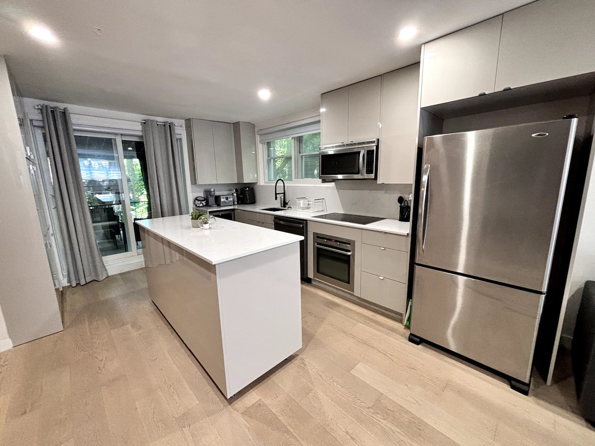 A modern kitchen is showcased, featuring sleek cabinetry, stainless steel appliances, and a spacious island with a white countertop. Natural light streams in through the window, complemented by soft gray curtains, creating a bright and functional cooking space.