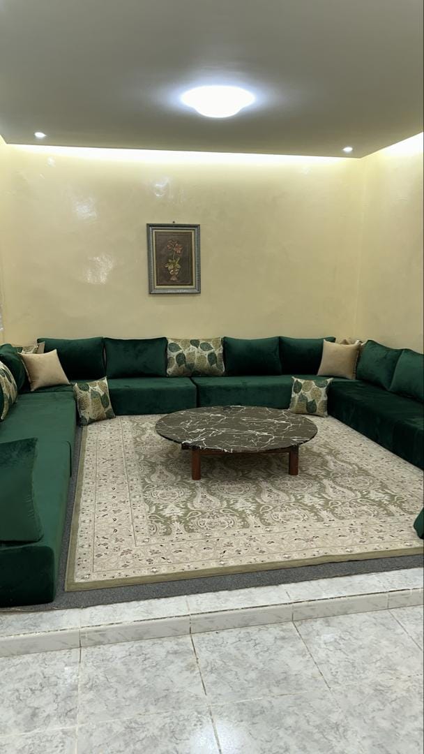 A spacious living area features a large L-shaped green sofa adorned with decorative pillows. The seating arrangement surrounds a round marble coffee table, positioned on an intricately patterned area rug that lies atop a light-colored floor.